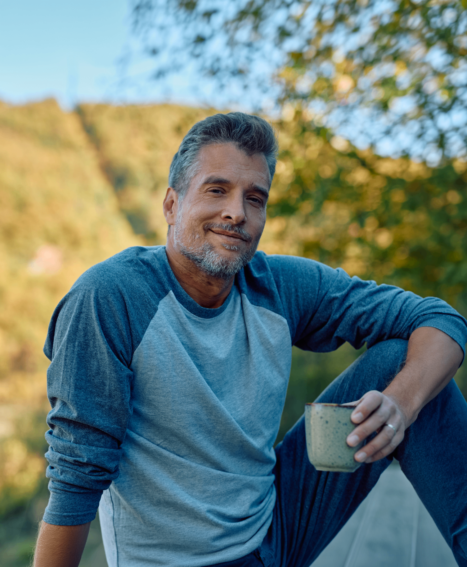 A calm, grounded adult outdoors with a warm smile, holding a cup
